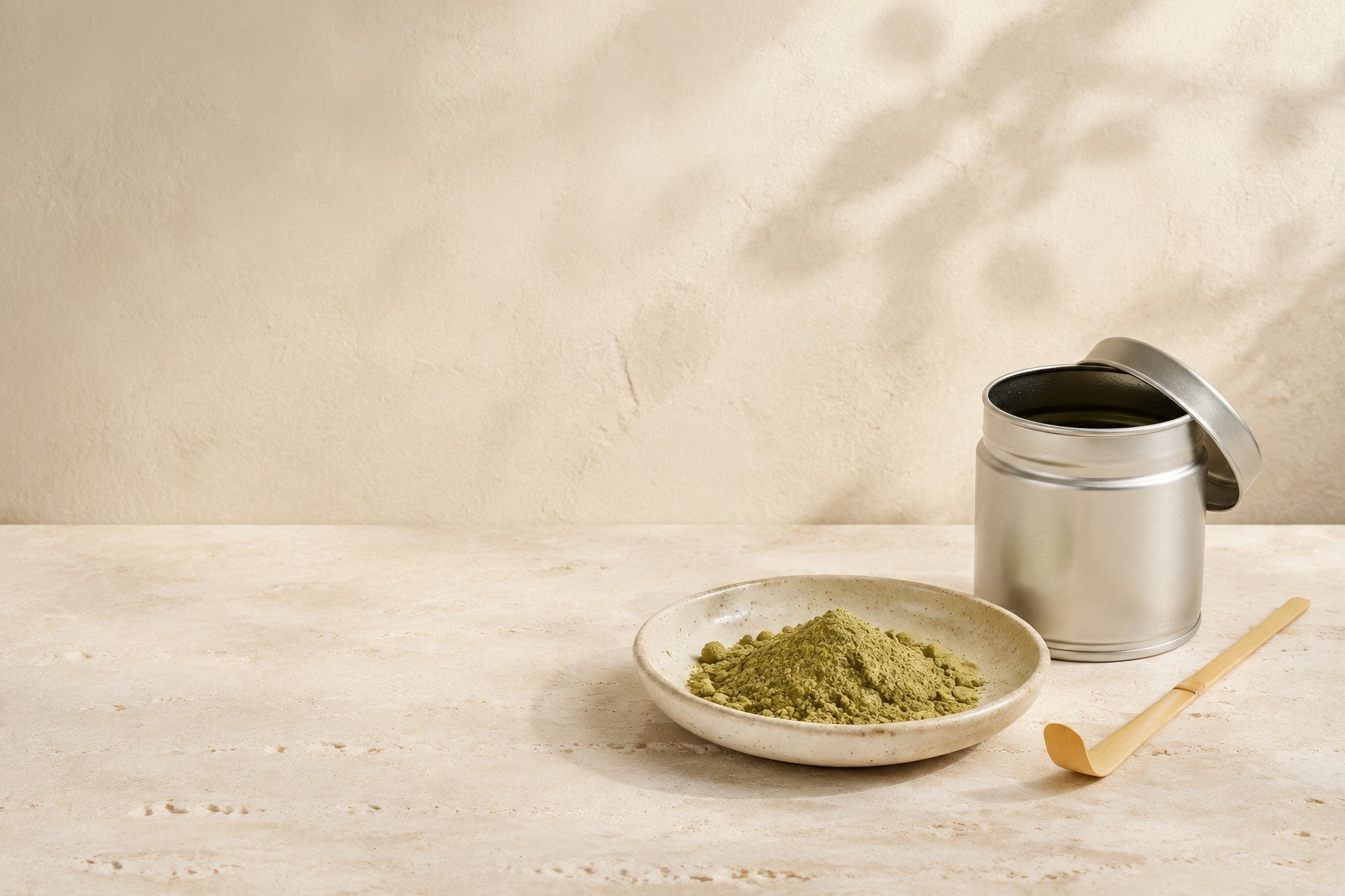 An open tin of matcha with its lid resting against the side, a shallow dish of dull olive-green powder and a bamboo scoop on a travertine surface, with dappled shadows on the wall behind