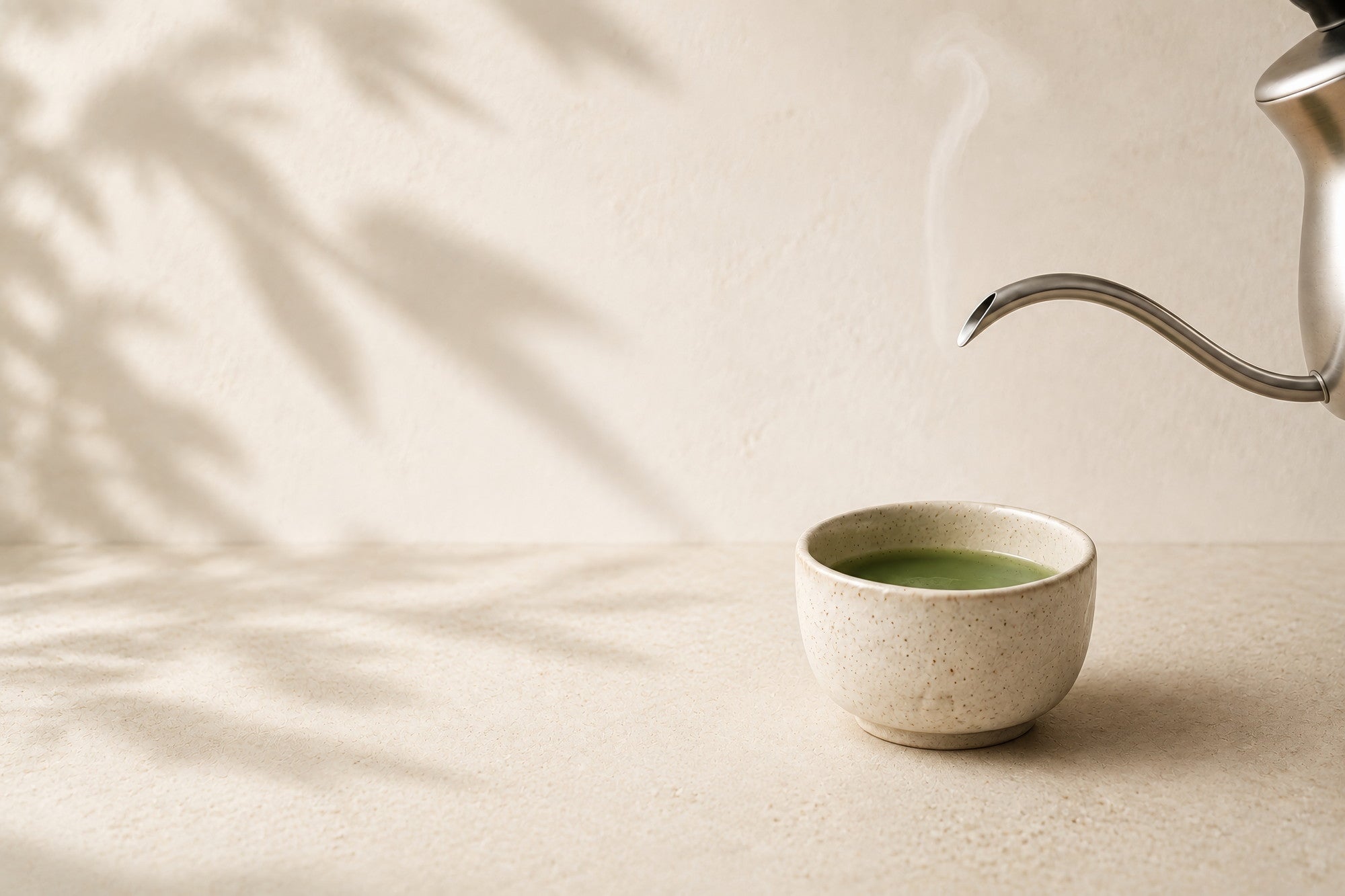 A gooseneck kettle poised above a small speckled ceramic cup of whisked matcha on a pale stone surface, steam rising, with soft leaf shadows on the wall behind