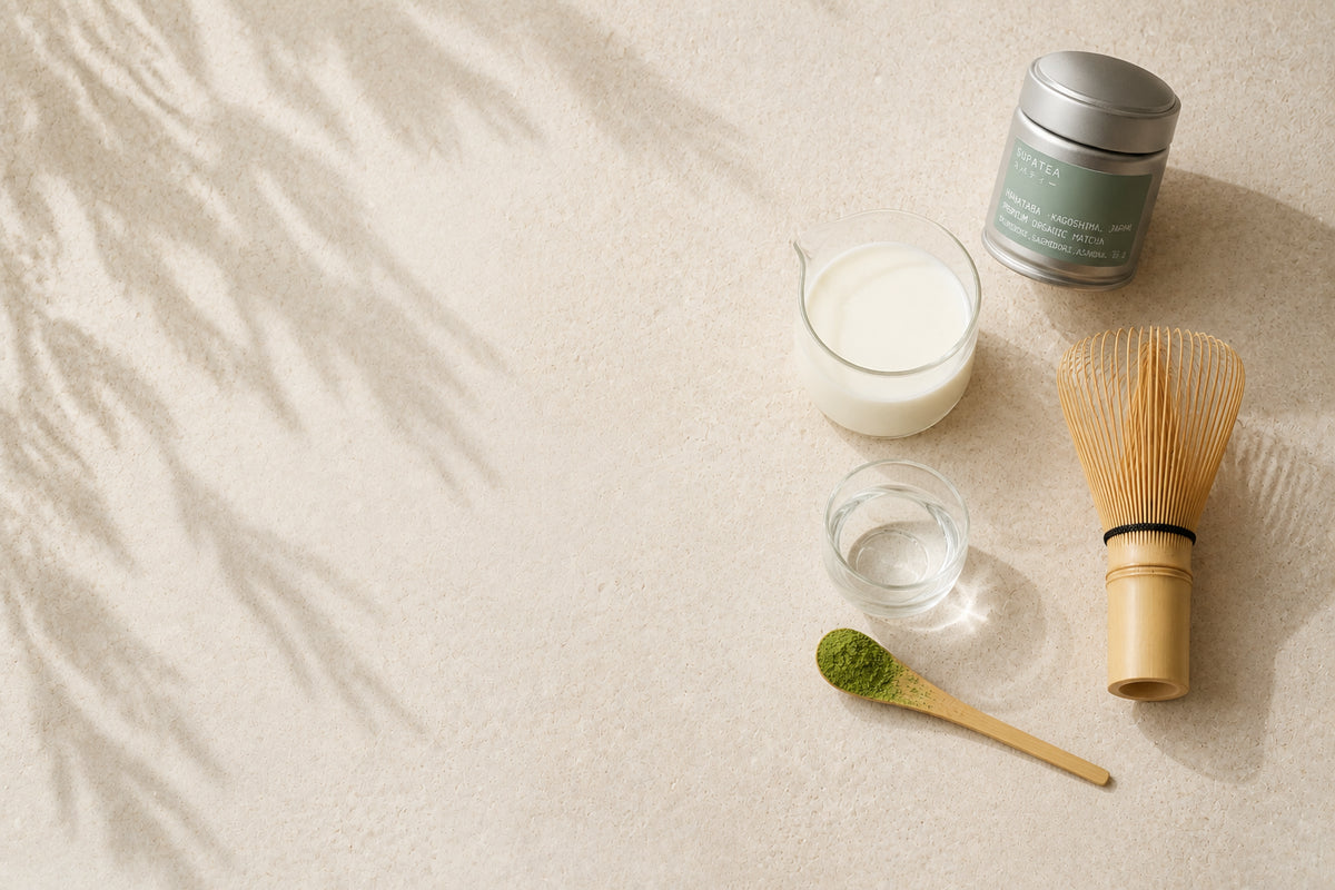 The ingredients for a hot matcha latte seen from above — a SUPATEA HANATABA tin, a jug of milk, a glass of water, a bamboo whisk and a wooden spoon of matcha powder