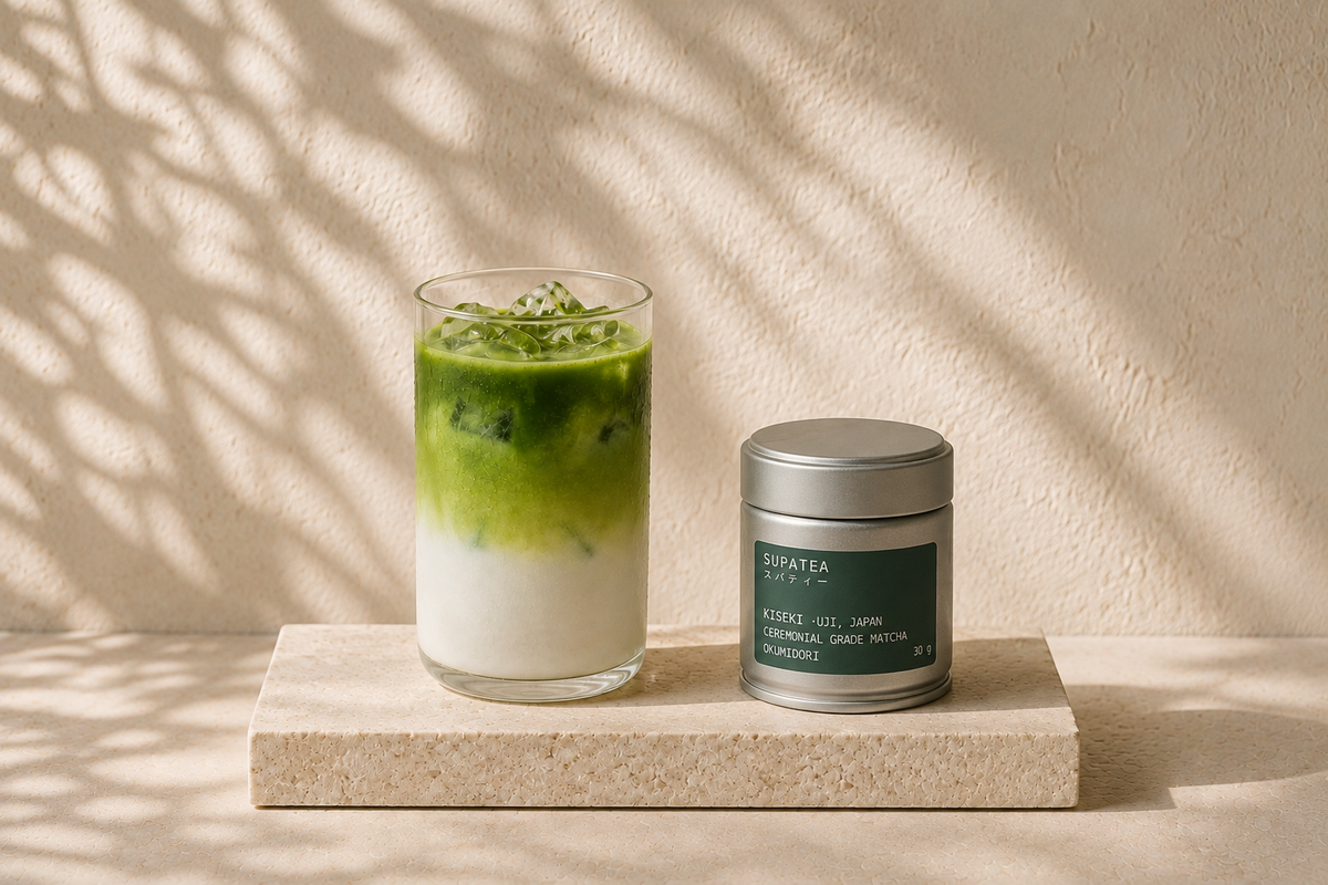 A tall glass of iced matcha latte, green layered over milk with ice, beside a SUPATEA KISEKI tin on a stone ledge