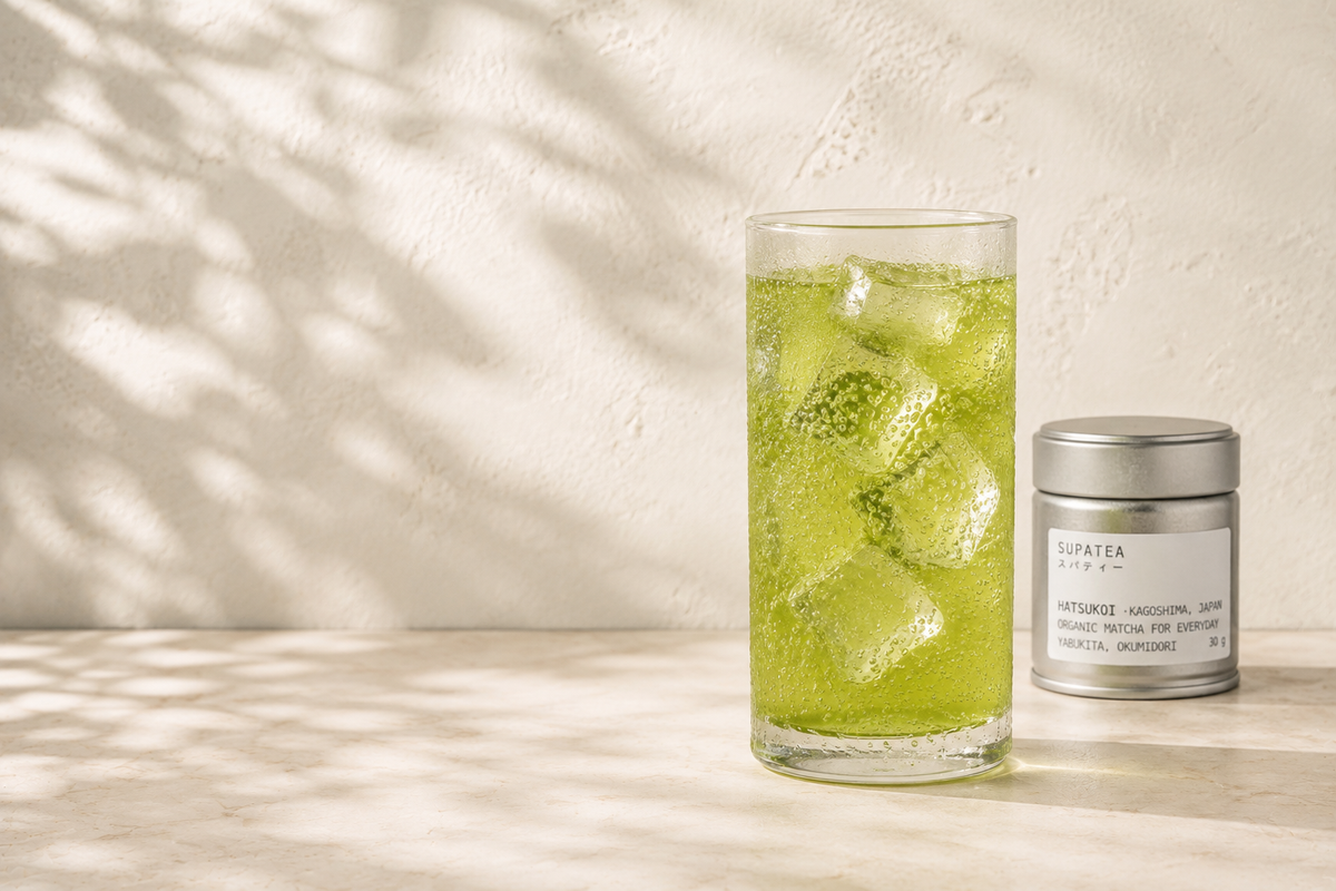 A tall glass of matcha and sparkling water over ice, bubbles rising, beside a SUPATEA HATSUKOI tin
