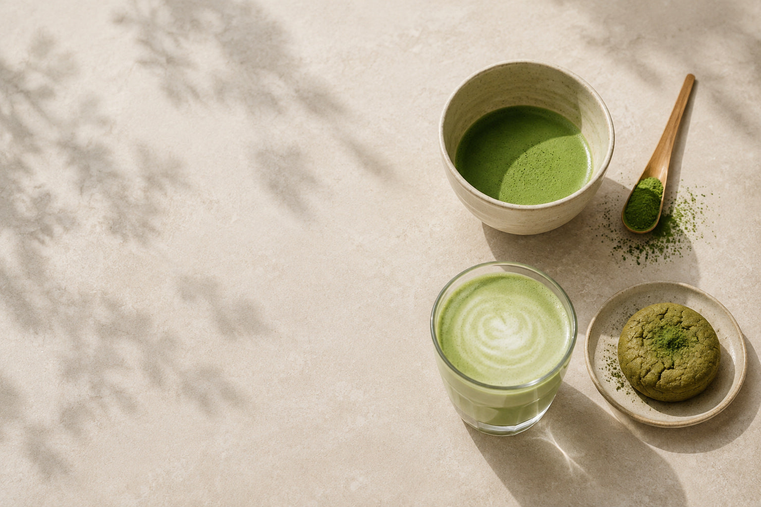 Seen from above: a bowl of whisked matcha, a wooden spoon holding loose powder, an iced matcha latte in a glass and a matcha cookie on a small plate, on a pale stone surface with leaf shadows