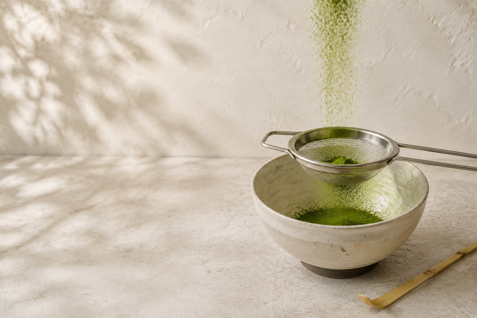 Bright green matcha falling in a fine stream through a mesh sieve into a pale ceramic bowl, with a bamboo whisk beside it and leaf shadows on the wall behind