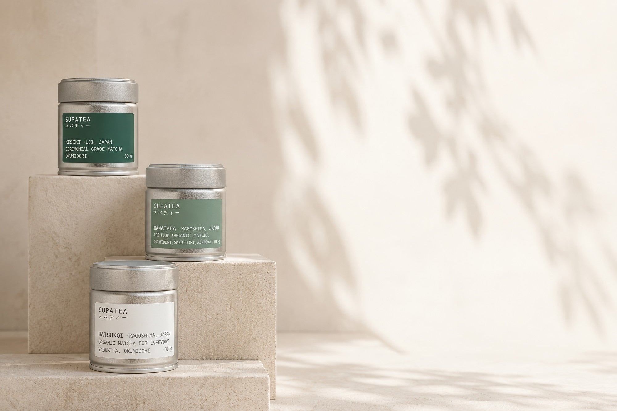 The three SUPATEA tins arranged on travertine blocks at different heights — KISEKI in deep green at the top, HANATABA in sage in the middle, HATSUKOI in white below — with dappled shadows on the wall behind